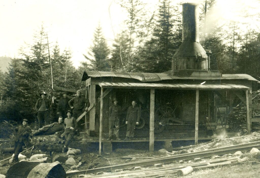 Eight men pose near of a stationary steam engine with a large cylindrical burner and smoke stack near a set of narrow gauge railroad tracks