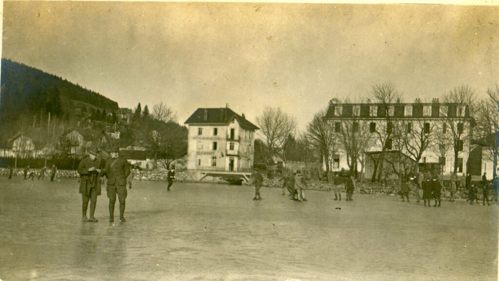 two soldiers pose on an ice skating rink.  in the background, children skate and two large buildings of a town are beyond that