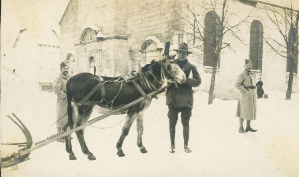 An American soldier posing with a donkey hitched to a sleigh.  two french soldiers look on smiling