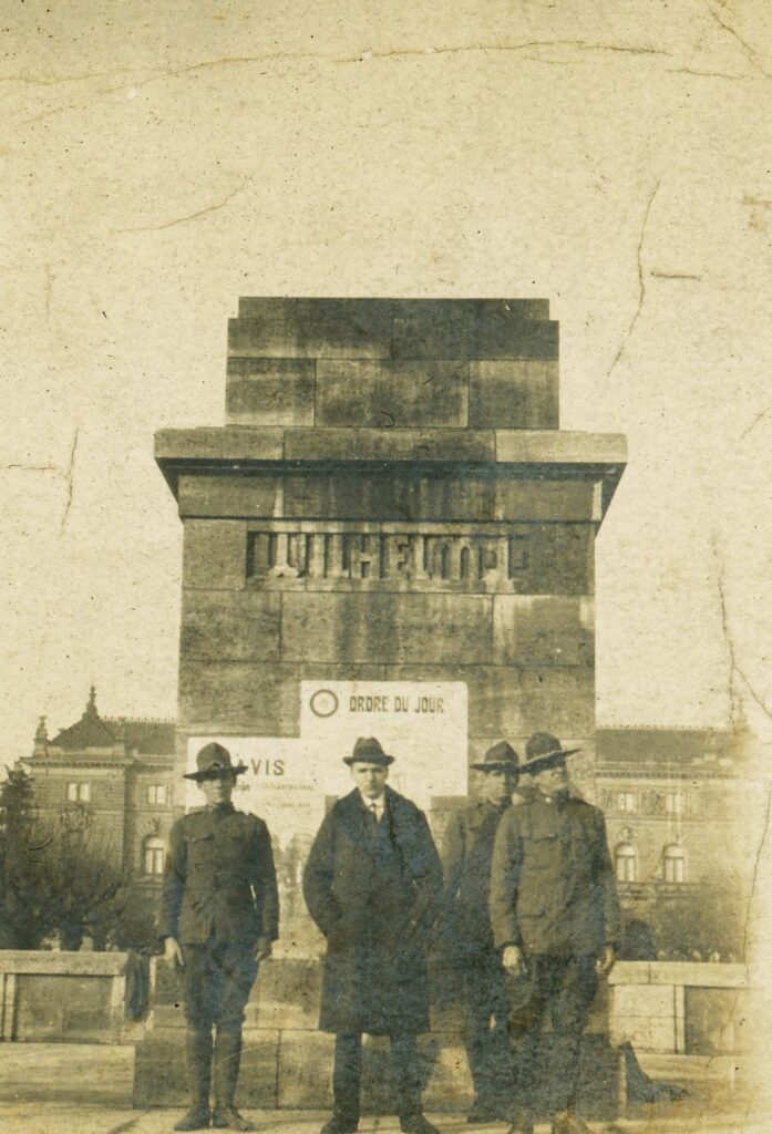 three men stand in front of an empty pedestal for a statue with the word "Wilhelm" engraved on it.