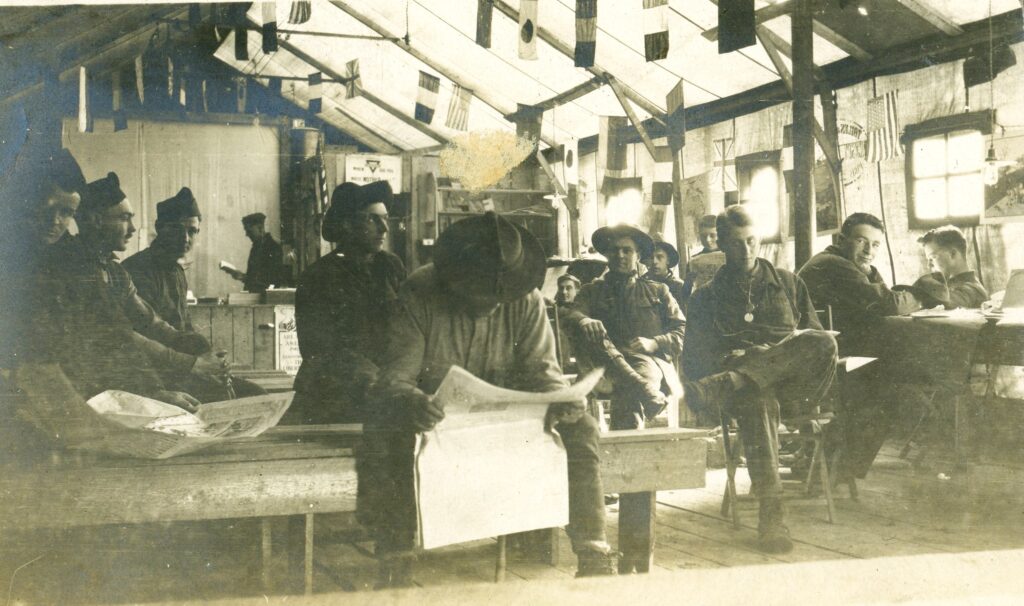 A group of young men sit inside a building under flag decorations.  Many of them are reading newspapers.