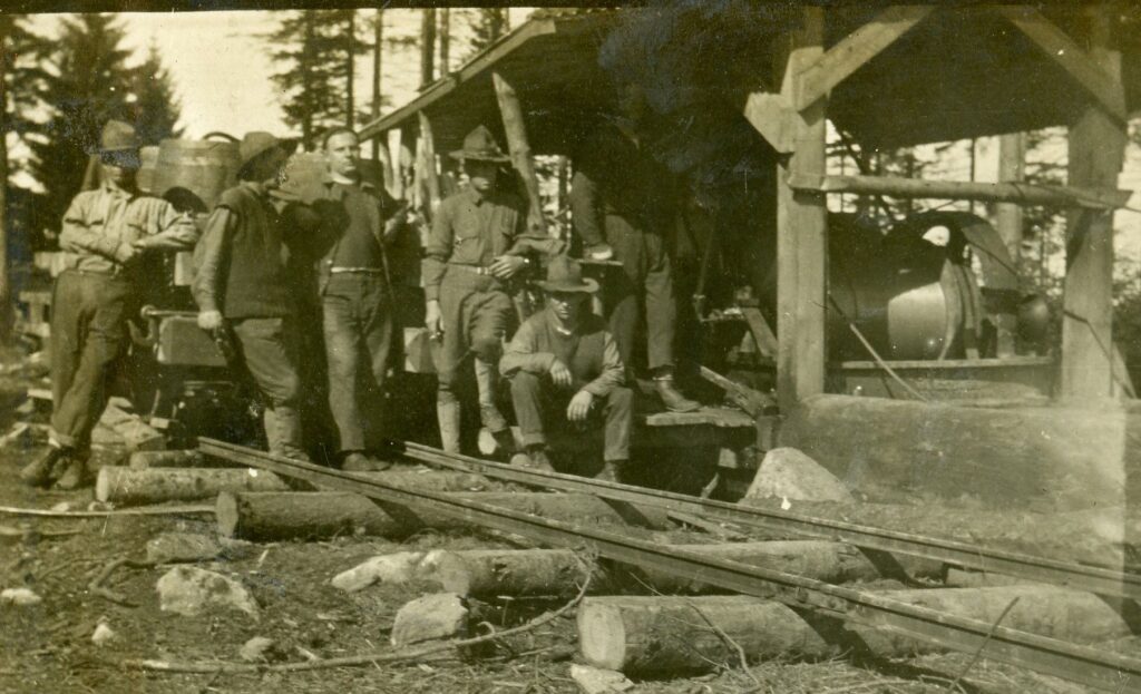 Six men in US Army uniforms pose near a shed with steam engine machinery inside