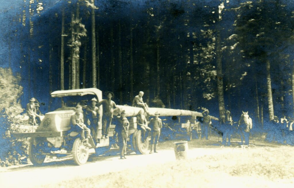 a group of men pose near a truck with the emblem of the 20th Engineers on the front. a very long log is loaded on the truck. a team of horses or mules stands to the right